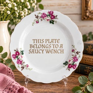 Puede incluir: Un plato de cerámica blanca con borde festoneado y acabado dorado, decorado con racimos de rosas rosas y blancas a lo largo del borde. El centro del plato muestra el texto dorado: THIS PLATE BELONGS TO A SAUCY WENCH, en una fuente decorativa.