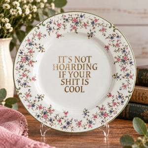 Puede incluir: Un plato de cerámica blanca con un borde floral de pequeñas flores rosas y azules y hojas verdes. El centro del plato muestra el texto en letras doradas: IT'S NOT HOARDING IF YOUR SHIT IS COOL. El plato se exhibe sobre un soporte transparente.