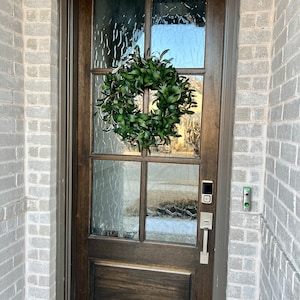 May include: A dark brown wooden front door with a glass pane and a green wreath. The door has a silver door handle and a silver keypad.