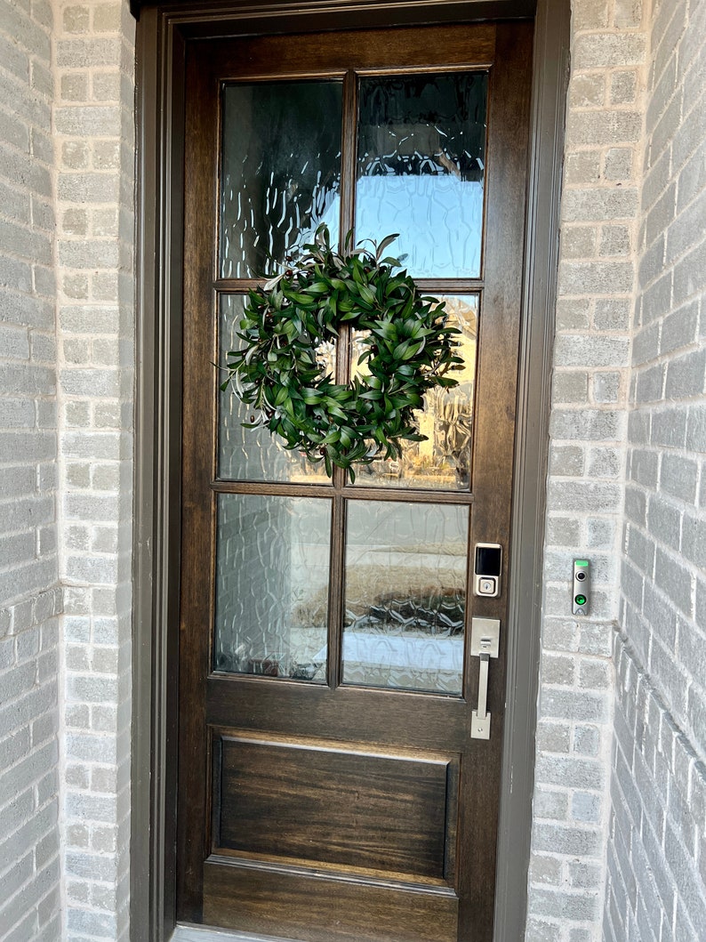 May include: A dark brown wooden front door with a glass pane and a green wreath. The door has a silver door handle and a silver keypad.
