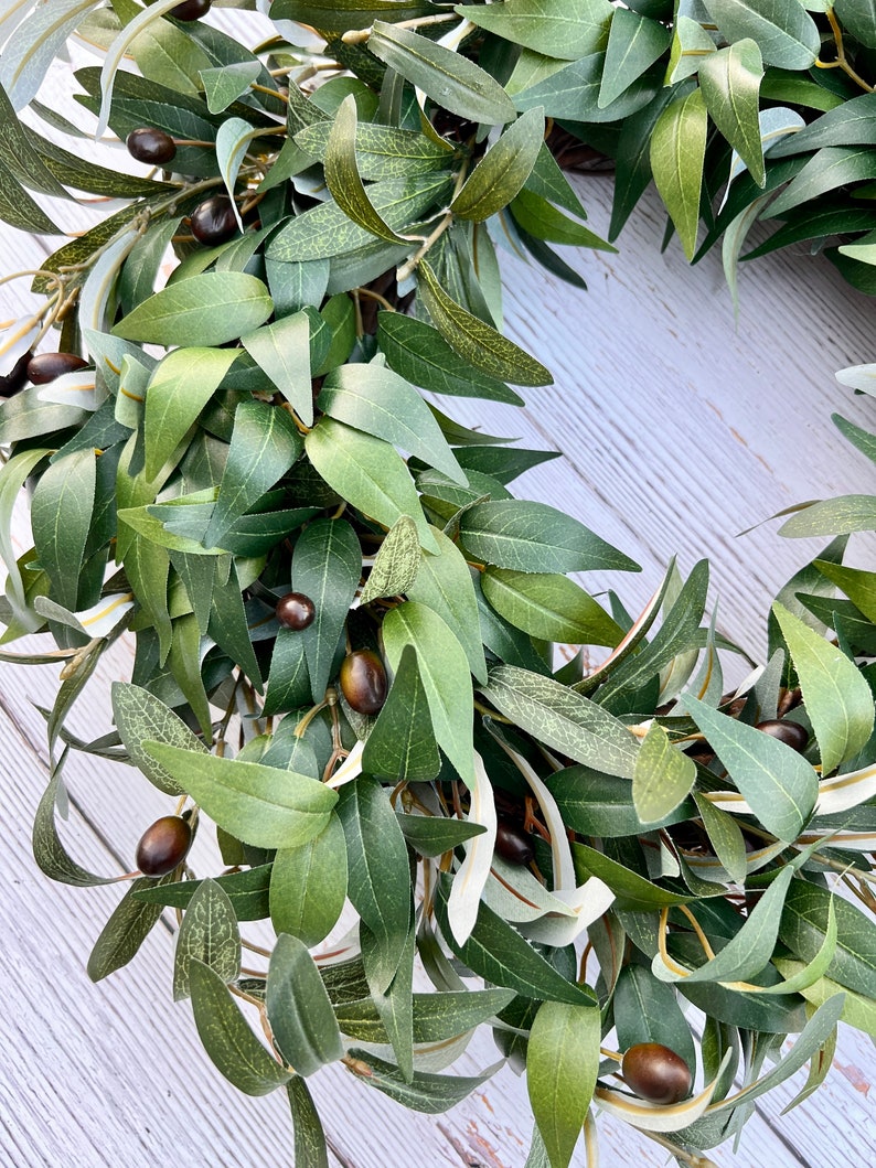 May include: A green olive branch wreath with brown olives. The wreath is made of artificial leaves and is perfect for adding a touch of nature to your home decor.