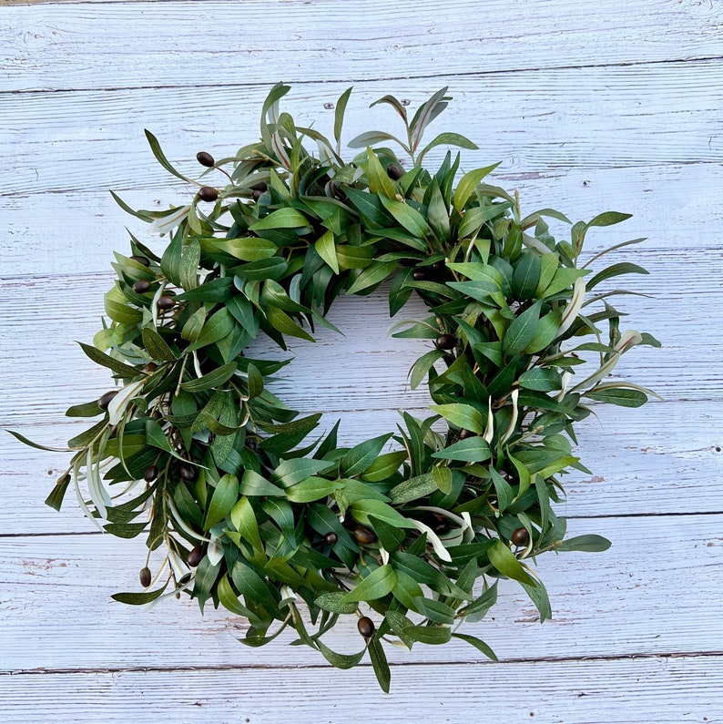 May include: A green olive branch wreath with brown olives. The wreath is made of natural materials and is perfect for adding a touch of nature to your home decor.