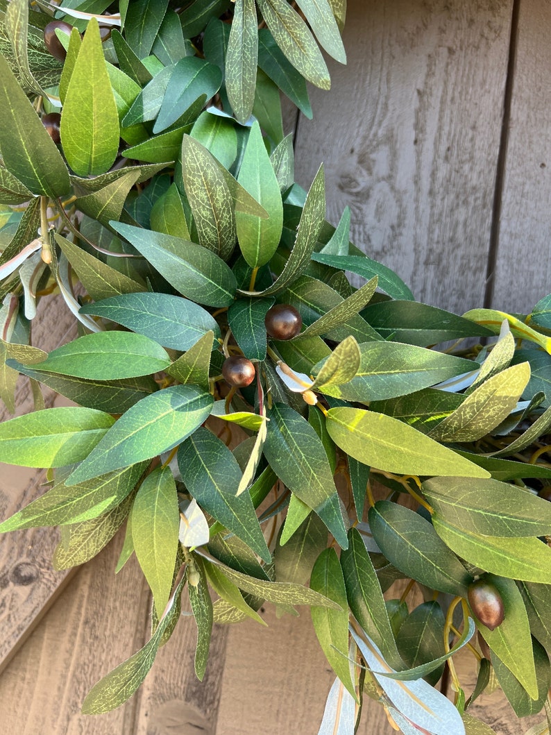 May include: A green olive branch wreath with brown olives. The wreath is made of artificial leaves and is perfect for adding a touch of nature to your home decor.