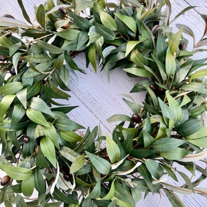May include: A green olive branch wreath with small, dark green olives. The wreath is made of artificial leaves and is perfect for adding a touch of nature to your home decor.