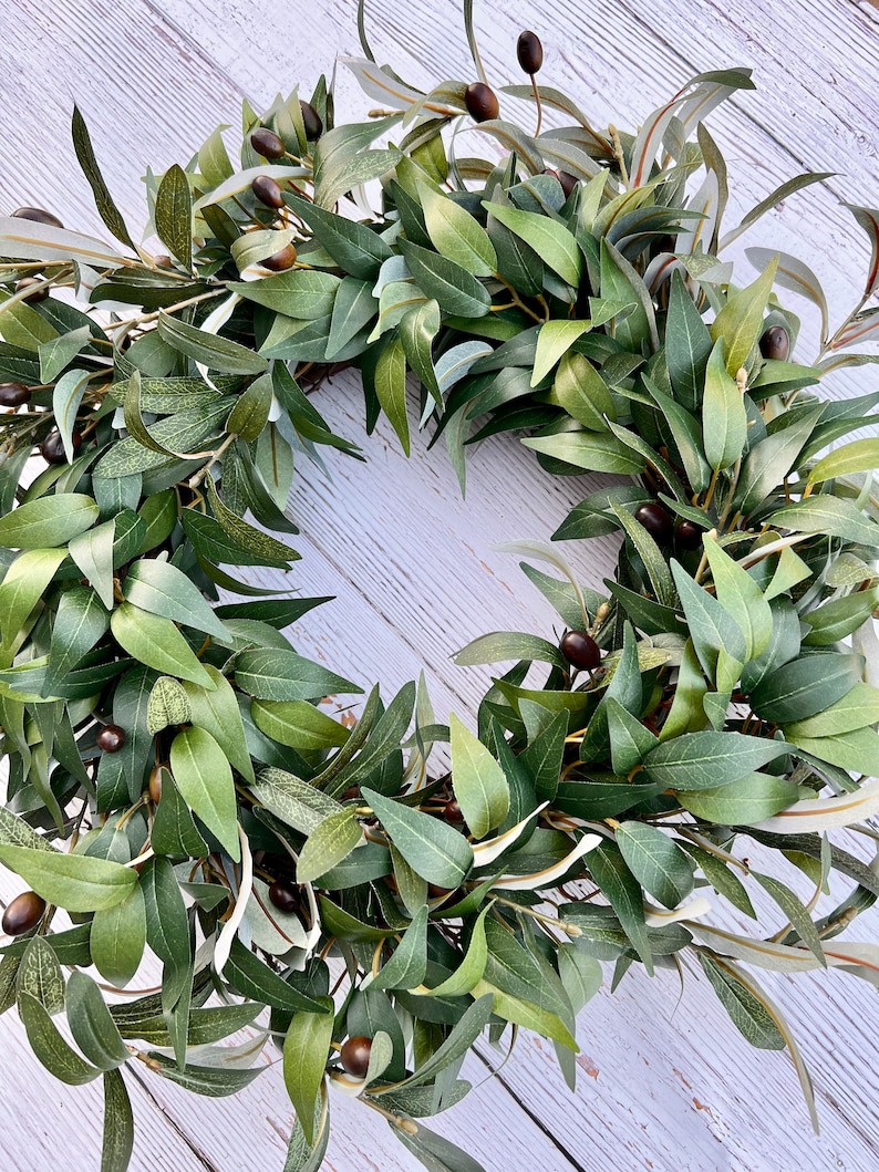 May include: A green olive branch wreath with small, dark green olives. The wreath is made of artificial leaves and is perfect for adding a touch of nature to your home decor.