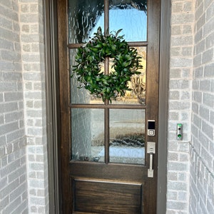 May include: A dark brown wooden front door with a wreath of green leaves hanging on it. The door has a glass panel with a textured pattern and a silver door handle. There is a silver doorbell and a security camera on the wall next to the door.