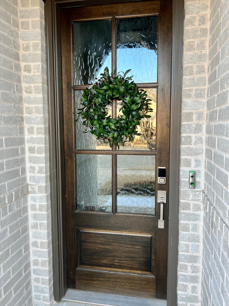 May include: A dark brown wooden front door with a wreath of green leaves hanging on it. The door has a glass panel with a textured pattern and a silver door handle. There is a silver doorbell and a security camera on the wall next to the door.