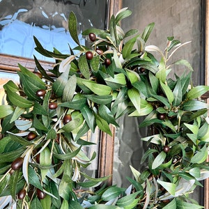 May include: A green olive branch wreath with brown olives. The wreath is hanging on a wooden door with a glass pane.