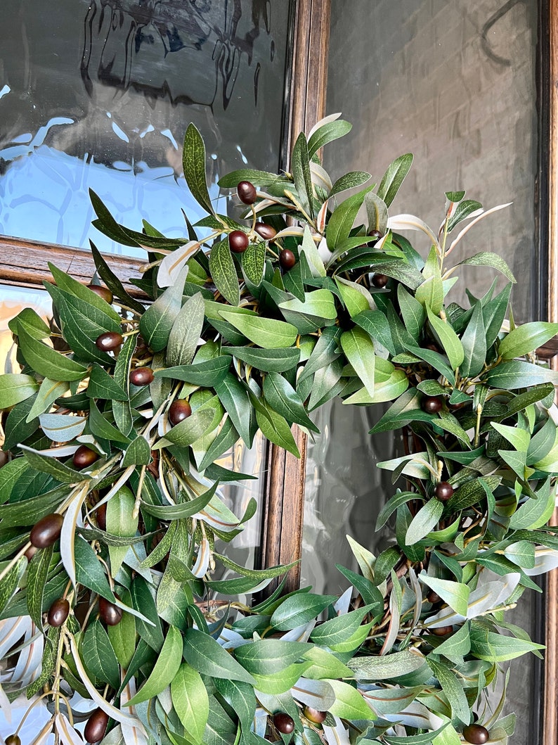 May include: A green olive branch wreath with brown olives. The wreath is hanging on a wooden door with a glass pane.