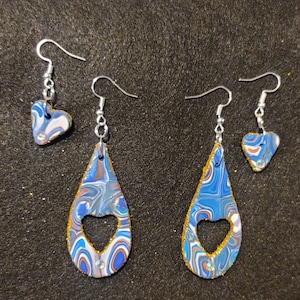 May include: A pair of dangle earrings with a blue and gold marbled design. Each earring features a heart-shaped cutout and a smaller heart charm.