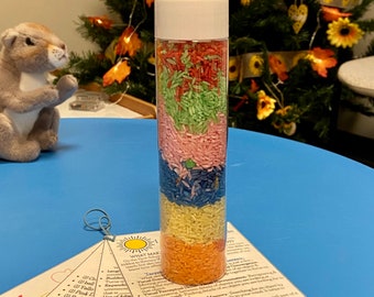 May include: A clear plastic bottle filled with layers of colored rice. The rice is red, green, pink, blue, yellow, and orange. The bottle is sitting on a blue surface with a white background. The bottle is labeled "What Makes This Bottle Special?" and lists several benefits of using a sensory bottle.