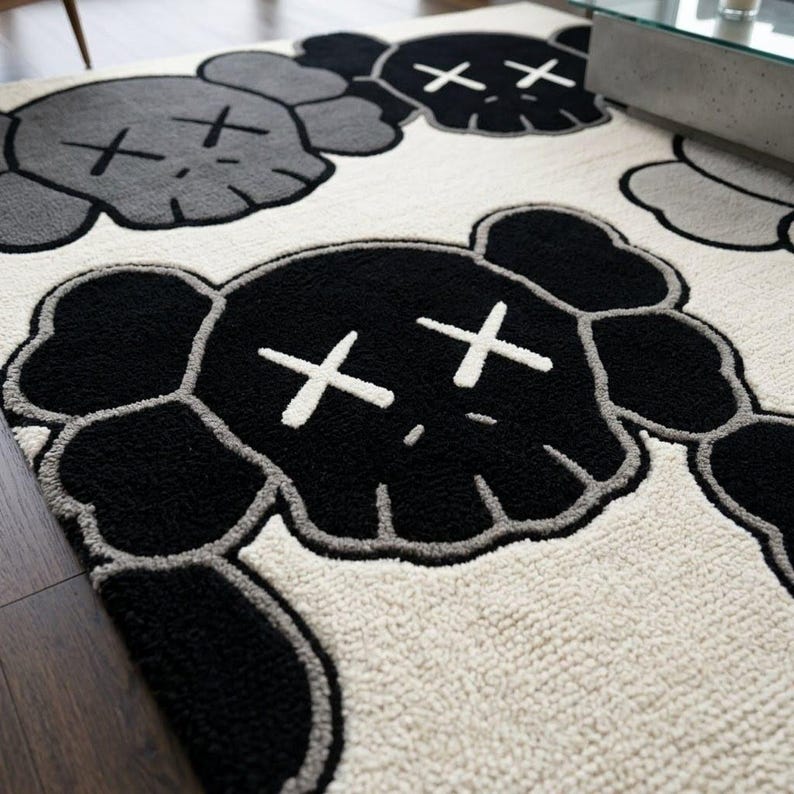 Close-up corner of a premium hand-tufted New Zealand wool rug featuring a black KAWS-inspired character head with white X eyes, showing the dense cream wool pile, deep black outlines, and high-low hand-carved textures on a dark hardwood floor.