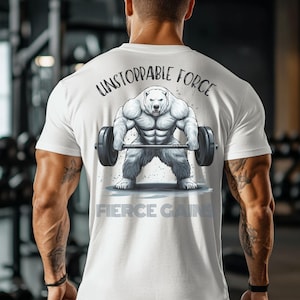 Può includere: Maglietta bianca con una grafica di un orso bianco che solleva un bilanciere. Il testo "Unstoppable Force" è sopra l'orso e "Fierce Gains" è sotto l'orso.