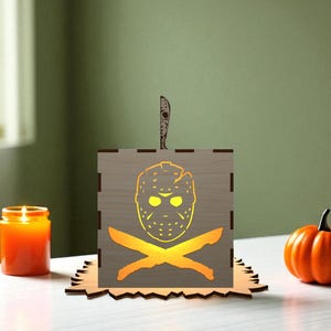 May include: A wooden Halloween-themed lantern with a cutout of a hockey mask and crossed knives, illuminated from within. An orange pumpkin and lit candle are nearby. The lantern is on a decorative wooden base.