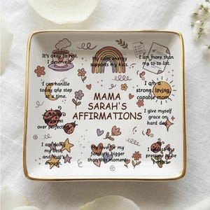 Könnte beinhalten: Eine quadratische Keramik-Schmuckschale mit goldenem Rand, mit Affirmationen und dem Text "MAMA SARAH'S AFFIRMATIONS". Die Schale ist mit farbenfrohen Illustrationen eines Regenbogens, Blumen und Marienkäfern verziert.