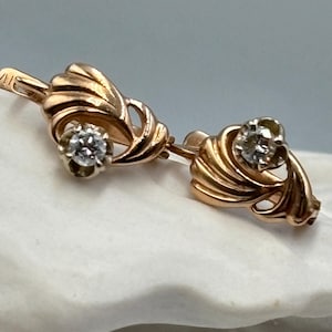 Könnte beinhalten: Ein Paar goldfarbene Ohrringe mit floralem Design. Jeder Ohrring hat einen funkelnden, rund geschliffenen Diamanten. Die Ohrringe werden auf einer weißen Oberfläche vor einem sanften blauen Hintergrund präsentiert. Die Ohrringe haben eine Vintage-Ästhetik.