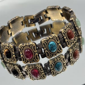 Op de afbeelding: Een vintage-stijl armband met een bronskleurige metalen ketting. Elke vierkante schakel heeft een sierlijk ontwerp en een kleurrijke edelsteen, waaronder rode, turquoise en bruine stenen. De armband wordt op een reflecterend oppervlak weergegeven.