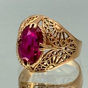 Peut inclure: Une bague dorée ornée d'une pierre précieuse ovale rouge foncé. La bague présente un motif filigrané complexe autour de la pierre et sur le corps. La pierre est maintenue par des griffes. La bague est présentée sur une surface réfléchissante.
