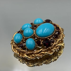Può includere: Un broche dorato con un gruppo di cabochon turchesi e strass rossi. Il broche è circondato da un design a corda intrecciata.