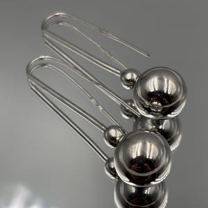 May include: A pair of silver-toned drop earrings featuring a long, slender wire hook that extends into a small bead and a larger, polished spherical ball at the end. The metal surface is highly reflective, showing a smooth, metallic finish.