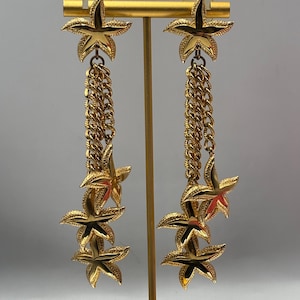 May include: A pair of long gold-tone dangle earrings featuring a top starfish stud connected to a textured chain link drop. Each earring displays two additional dangling starfish charms at the base, creating a layered, multi-dimensional gold design.