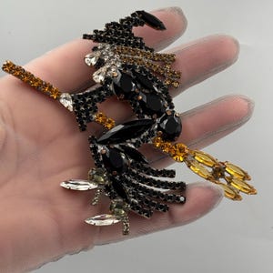 Puede incluir: Un broche con forma de bruja decorado con pedrería en tonos negros, transparentes y ámbar. El diseño incluye un sombrero puntiagudo, una capa fluida y una escoba. Las piedras están engastadas en una estructura metálica, creando un acabado brillante.