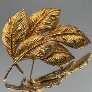 May include: A gold-toned metal brooch shaped like a cluster of three textured leaves. The surface of the leaves features intricate, raised vein details and a metallic finish that catches the light, creating a realistic appearance of foliage.
