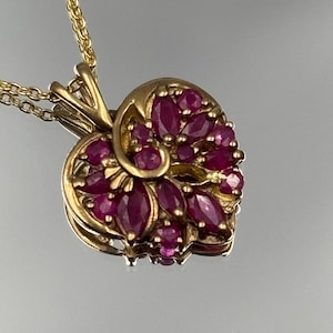 Può includere: Una collana con pendente in oro dal design a forma di cuore, decorata con molteplici rubini rosso scuro a taglio marquise e rotondo. La montatura in oro include un accento metallico a spirale che si curva al centro del cuore, su una catena d'oro.