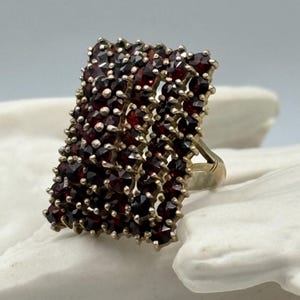 Puede incluir: Un anillo de plata con una cara rectangular engastada con un grupo de pequeñas piedras de granate rojo oscuro.