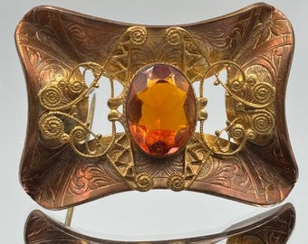 Op de afbeelding: Een antieke broche met een koperkleurige basis en gouden filigraanaccenten. De broche heeft een ovale, amberkleurige edelsteen in het midden. Er zijn ingewikkelde bloempatronen in het koper geëtst, wat bijdraagt aan de vintage charme.