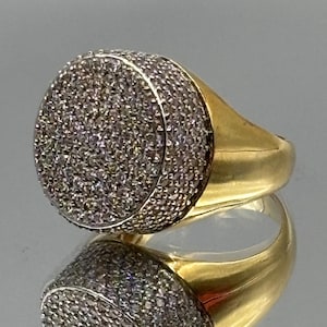 Puede incluir: Un anillo de oro con una cara circular grande densamente incrustada con numerosas piedras pequeñas, claras y brillantes. La banda es de oro liso y pulido, conectada a la montura circular que está completamente cubierta de piedras.