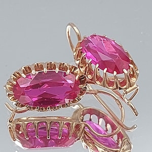 May include: A pair of gold earrings featuring pink oval-shaped gemstones set in a delicate gold frame. The earrings have a simple, elegant design.