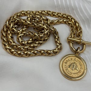 May include: A gold-coloured necklace with a thick chain and a circular pendant. The pendant features an embossed bee design and the words "JOAN RIVERS COLLECTION". The necklace has a toggle clasp.