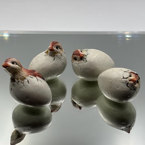 May include: Four small, white ceramic eggs with cracked shells, each containing a detailed, painted baby bird. The birds have brown and red markings. The eggs are reflected on a shiny surface, creating a symmetrical image.