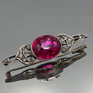 May include: A silver bar brooch with a large, oval, magenta gemstone. The gemstone is set in silver and surrounded by smaller, clear gemstones. The brooch has an ornate, Art Deco design. The brooch is approximately 6 cm in length.