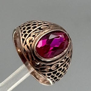 Può includere: Un anello color argento con una gemma color magenta vibrante dal taglio ovale incastonata al centro. La fascia presenta un intricato motivo in metallo traforato che ricorda un design a scaglie o a reticolo sui lati dell'anello.