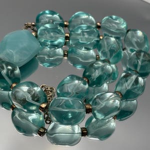Peut inclure: Un bracelet composé de perles en verre translucide bleu clair, arrondies et dotées d'une texture interne ondulée, séparées par de petites entretoises métalliques. Il comprend une perle opaque bleu clair plus grande et une chaîne en métal argenté.