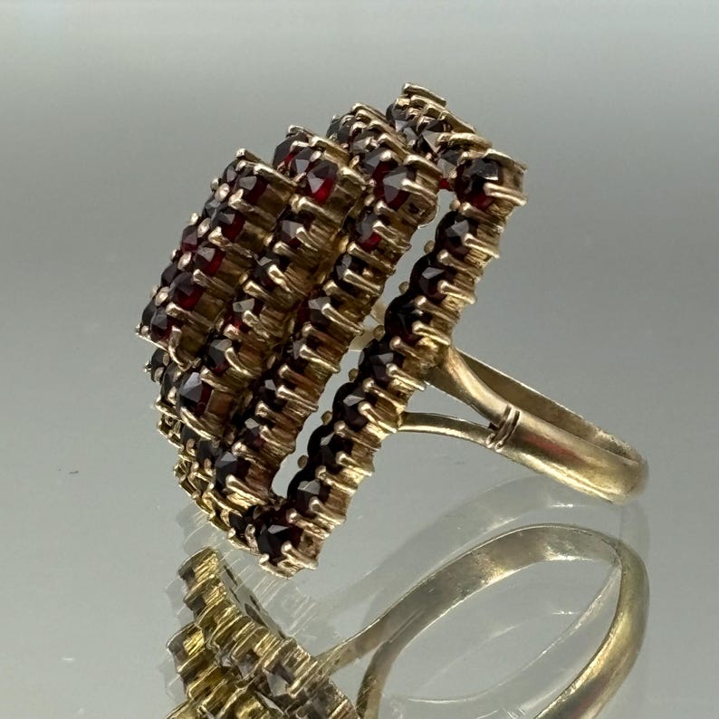 Op de afbeelding: Een gouden ring met een grote, vierkante cluster van rode granaten. De ring heeft een eenvoudige band en de granaten zijn in een aflopend patroon gezet.