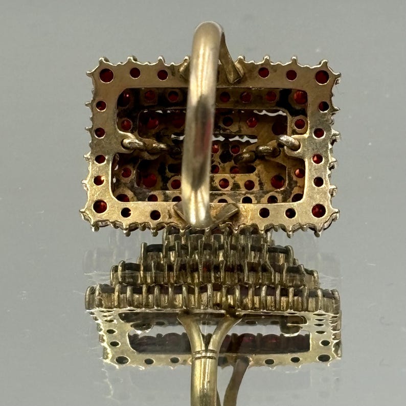 Op de afbeelding: Een gouden ring met een rechthoekige zetting gevuld met rode edelstenen. De ring is bezet met een groot aantal kleine, ronde stenen.