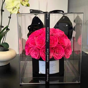 May include: A clear acrylic box contains a bouquet of bright pink roses. The roses are arranged in a black container, with a black ribbon printed with "Forever Love." A small white card is visible on the front of the container.