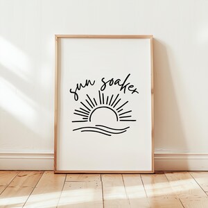 Puede incluir: Una impresión en blanco y negro con un dibujo de línea simple de un sol poniéndose sobre las olas. El texto "sun soaker" está escrito sobre el sol.