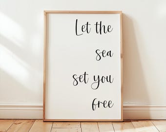May include: A framed print with the words "Let the sea set you free" in black script. The print has a white background and a light brown wooden frame. It is leaning against a white wall on a wooden floor.