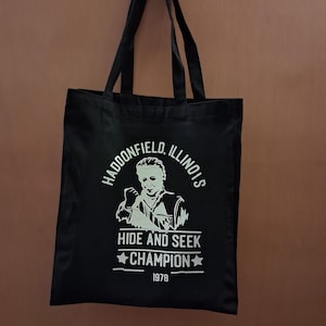 Puede incluir: Bolso tote negro con texto e imagen gráficos en blanco. El texto dice "HADDONFIELD, ILLINOIS" encima de una imagen de una persona. Debajo de la imagen está el texto "HIDE AND SEEK CHAMPION 1978". El bolso tiene dos asas negras.