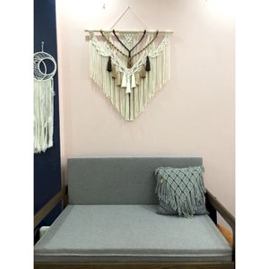 May include: A white macrame wall hanging with brown and beige tassels, hanging on a pink wall above a gray couch with a blue and white macrame pillow.