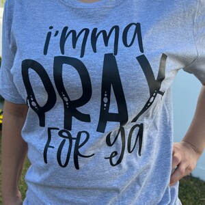Puede incluir: Camiseta gris claro con la frase "i'mma PRAY FOR ya" en letras negras grandes. La camiseta de manga corta parece estar hecha de un material suave y es de estilo casual.