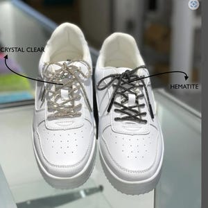 Puede incluir: Dos zapatillas blancas con cordones decorativos. Un par tiene cordones cristalinos y el otro tiene cordones de hematita. Las zapatillas tienen un diseño clásico con suela blanca y puntera perforada.
