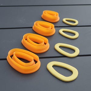 Peut inclure: Un ensemble de quatre emporte-pièces ovales en plastique orange disposés en colonne verticale, accompagnés de quatre découpes ovales jaunes en pâte ou argile disposées en colonne parallèle sur la droite des emporte-pièces.