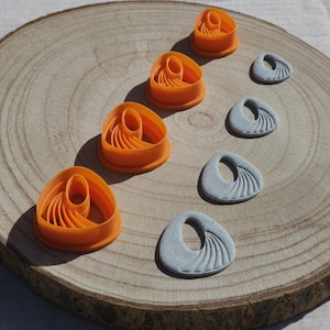 May include: Four orange plastic cutters with a triangular shape and internal curved ribbing details, accompanied by four corresponding light gray speckled clay shapes that feature a matching embossed fan-like pattern on their surfaces.