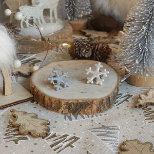 Peut inclure: Deux ornements en forme de flocon de neige, un argenté pailleté et un blanc, sont posés sur une tranche de bois. La scène comprend des sapins miniatures, des pommes de pin et des décorations en forme de feuilles, le tout sur une nappe à motifs festifs.
