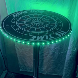 Könnte beinhalten: Runder, schwarzer Dartboard-Tisch mit grünem LED-Lichtrand. Das Tischdesign zeigt eine Dartscheibe mit Zahlen und den Texten "IT'S TIME" und "FOR DART". Er steht auf einem silbernen Metallfuß.